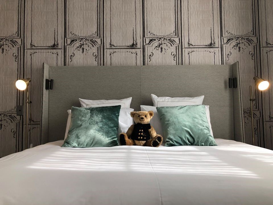 Teddy bear wearing jacket sitting on hotel bed between pillows
