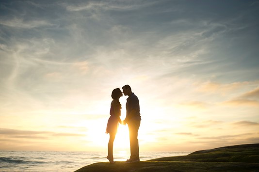 Photo of two people about to kiss with sunset and the ocean in the background.